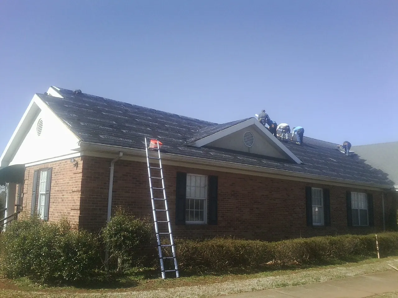 Crew tearing shingles off a brick ranch roof with a ladder set — Emergency Roof Repair work by our crew in Anthon