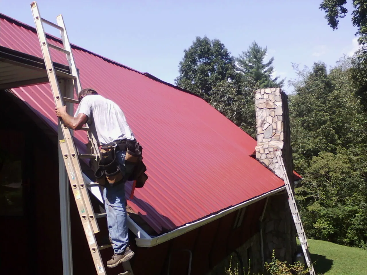 Roofer on a ladder beside a red metal roof and stone chimney — Soffit & Fascia Repair by Roofing Brothers in Anthon